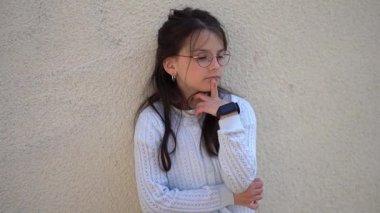 Kırmızı gözlüklü güzel bir kızın düşünceli bir şekilde duvarın yanında durduğu yatay 4K video. Akıllı çocuk konsepti