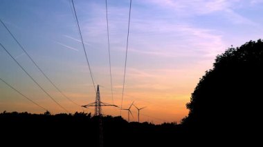İki rüzgar türbini, yüksek voltajlı bir elektrik hattının yanında turuncu-mavi günbatımında ufukta dönüyor. Yenilenebilir enerji ve kırsal arazinin 4K eş zamanlı videosu.