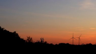 İki rüzgar türbini birbirine yakın turuncu-mavi bir günbatımına karşı dönüyor. Yenilenebilir enerji kavramı. 4k gerçek zamanlı video