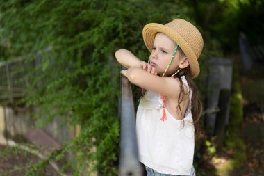 Hasır şapkalı küçük kız, yaz parkındaki korkuluklara dayanan mesafeye endişeli bir şekilde bakıyor. Çocukların boş vakitleri ve duyguları kavramı