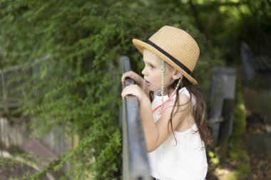 Hasır şapkalı küçük kız, yaz parkındaki korkuluklara dayanan mesafeye endişeli bir şekilde bakıyor. Çocukların boş vakitleri ve duyguları kavramı