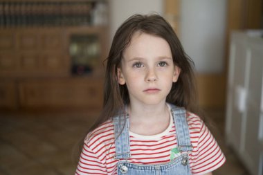 Odanın ortasında kot tulumlu şirin küçük bir kız duruyor. Arka plan bulanık. Çocukluk duyguları kavramı