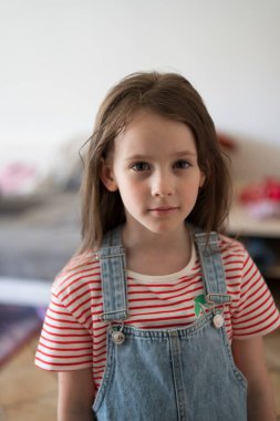 Odanın ortasında kot tulumlu şirin küçük bir kız duruyor. Arka plan bulanık. Çocukluk duyguları kavramı