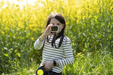Uzun saçlı, gözlüklü ve çizgili tişörtlü güzel bir kız baharda sarı kolza tohumu tarlasının önünde fotoğraf çekiyor. Çocukların yaratıcılığı ve rahatlığı kavramı