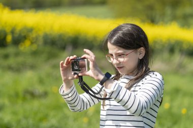 Uzun saçlı, gözlüklü ve çizgili tişörtlü güzel bir kız baharda sarı kolza tohumu tarlasının önünde fotoğraf çekiyor. Çocukların yaratıcılığı ve rahatlığı kavramı