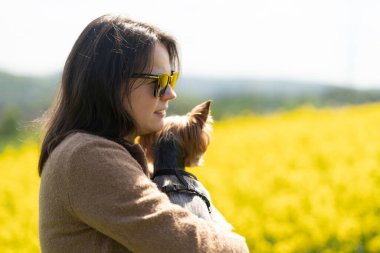 Kollarında küçük bir Yorkshire teriyeri olan güneş gözlüklü ve kazaklı güzel bir kız, sarı bir ırz tohumları tarlasının ortasında neşeli bir şekilde poz veriyor. Bir evcil hayvanla arkadaşlık kavramı