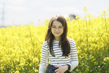 Gözlüklü ve çizgili tişörtlü şirin kız sarı kolza tohumu tarlasının önünde gülümsüyor. Çocukların duyguları ve boş vakitleri kavramı