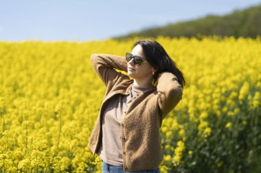 Güneş gözlüklü, tişörtlü ve süveterli güzel bir kız sarı bir kolza tohumu tarlasında neşeli bir şekilde poz veriyor. Duygular ve eğlence kavramı.