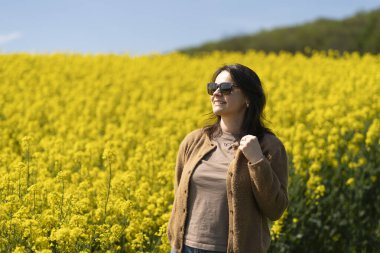 Güneş gözlüklü, tişörtlü ve süveterli güzel bir kız sarı bir kolza tohumu tarlasında neşeli bir şekilde poz veriyor. Duygular ve eğlence kavramı.