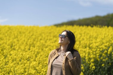 Güneş gözlüklü, tişörtlü ve süveterli güzel bir kız sarı bir kolza tohumu tarlasında neşeli bir şekilde poz veriyor. Duygular ve eğlence kavramı.