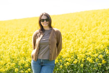 Güneş gözlüklü, tişörtlü ve süveterli güzel bir kız sarı bir kolza tohumu tarlasında neşeli bir şekilde poz veriyor. Duygular ve eğlence kavramı.
