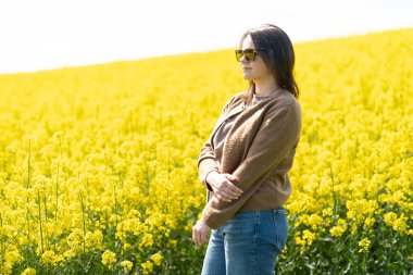 Güneş gözlüklü, tişörtlü ve süveterli güzel bir kız sarı bir kolza tohumu tarlasında neşeli bir şekilde poz veriyor. Duygular ve eğlence kavramı.
