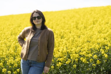 Güneş gözlüklü, tişörtlü ve süveterli güzel bir kız sarı bir kolza tohumu tarlasında neşeli bir şekilde poz veriyor. Duygular ve eğlence kavramı.