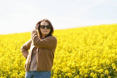 Güneş gözlüklü, tişörtlü ve süveterli güzel bir kız sarı bir kolza tohumu tarlasında neşeli bir şekilde poz veriyor. Duygular ve eğlence kavramı.