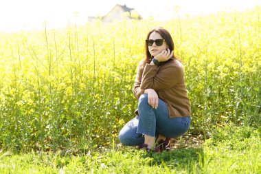 Güneş gözlüklü, tişörtlü ve süveterli güzel bir kız sarı bir kolza tohumu tarlasında neşeli bir şekilde poz veriyor. Duygular ve eğlence kavramı.