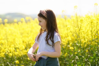 Beyaz tişörtlü ve kemerine kazak bağlanmış neşeli bir kız sarı bir kolza tohumu tarlasının ortasında neşeli bir şekilde duruyor. Çocukların duyguları ve boş vakitleri kavramı.