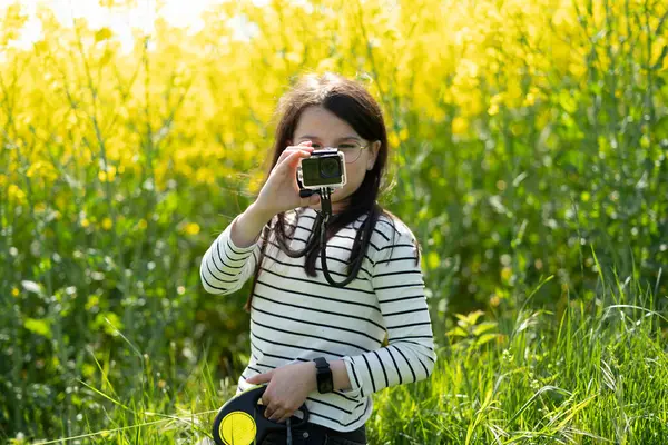Uzun saçlı, gözlüklü ve çizgili tişörtlü güzel bir kız baharda sarı kolza tohumu tarlasının önünde fotoğraf çekiyor. Çocukların yaratıcılığı ve rahatlığı kavramı