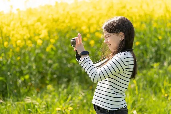 Uzun saçlı, gözlüklü ve çizgili tişörtlü güzel bir kız baharda sarı kolza tohumu tarlasının önünde fotoğraf çekiyor. Çocukların yaratıcılığı ve rahatlığı kavramı