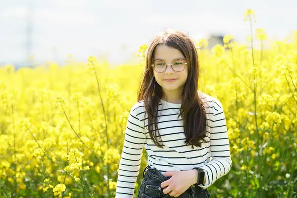 Gözlüklü ve çizgili tişörtlü şirin kız sarı kolza tohumu tarlasının önünde gülümsüyor. Çocukların duyguları ve boş vakitleri kavramı