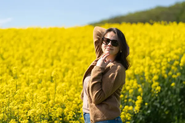 Güneş gözlüklü, tişörtlü ve süveterli güzel bir kız sarı bir kolza tohumu tarlasında neşeli bir şekilde poz veriyor. Duygular ve eğlence kavramı.