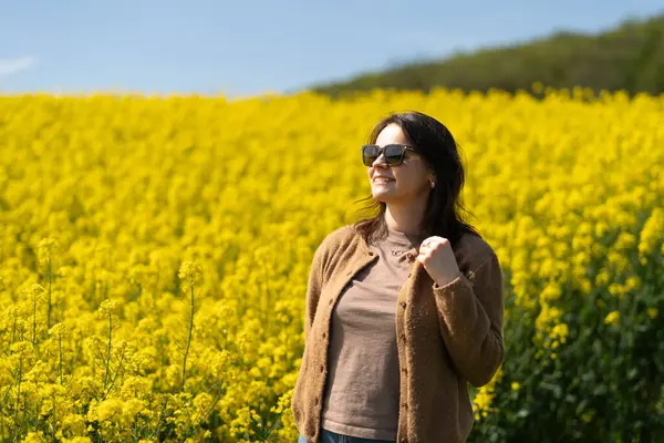 Güneş gözlüklü, tişörtlü ve süveterli güzel bir kız sarı bir kolza tohumu tarlasında neşeli bir şekilde poz veriyor. Duygular ve eğlence kavramı.