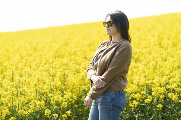 Güneş gözlüklü, tişörtlü ve süveterli güzel bir kız sarı bir kolza tohumu tarlasında neşeli bir şekilde poz veriyor. Duygular ve eğlence kavramı.