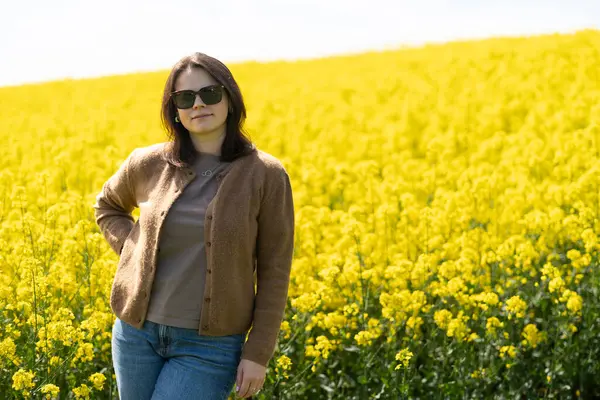 Güneş gözlüklü, tişörtlü ve süveterli güzel bir kız sarı bir kolza tohumu tarlasında neşeli bir şekilde poz veriyor. Duygular ve eğlence kavramı.