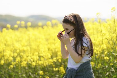 Beyaz tişörtlü, büyük güneş gözlüklü ve kemerinde süveteri olan neşeli bir kız sarı bir ırz tohumu tarlasının ortasında neşeli bir şekilde duruyor. Çocukların duyguları ve boş vakitleri kavramı.
