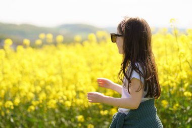 Beyaz tişörtlü, büyük güneş gözlüklü ve kemerinde süveteri olan neşeli bir kız sarı bir ırz tohumu tarlasının ortasında neşeli bir şekilde duruyor. Çocukların duyguları ve boş vakitleri kavramı.