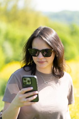 Güneş gözlüklü güzel, siyah saçlı bir kız ve elinde akıllı bir tişörtle sarı bir ırz tohumları tarlasının önünde fotoğraf çekiyor. Dijital yaşam ve doğa kavramı