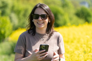 Güneş gözlüklü güzel, siyah saçlı bir kız ve elinde akıllı bir tişörtle sarı bir ırz tohumları tarlasının önünde fotoğraf çekiyor. Dijital yaşam ve doğa kavramı