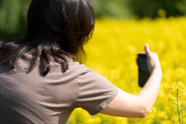 Güneş gözlüklü güzel, siyah saçlı bir kız ve elinde akıllı bir tişörtle sarı bir ırz tohumları tarlasının önünde fotoğraf çekiyor. Dijital yaşam ve doğa kavramı