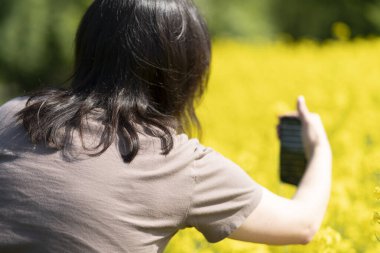 Güneş gözlüklü güzel, siyah saçlı bir kız ve elinde akıllı bir tişörtle sarı bir ırz tohumları tarlasının önünde fotoğraf çekiyor. Dijital yaşam ve doğa kavramı