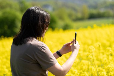 Güneş gözlüklü güzel, siyah saçlı bir kız ve elinde akıllı bir tişörtle sarı bir ırz tohumları tarlasının önünde fotoğraf çekiyor. Dijital yaşam ve doğa kavramı