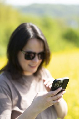 Güneş gözlüklü güzel, siyah saçlı bir kız ve elinde akıllı bir tişörtle sarı bir ırz tohumları tarlasının önünde fotoğraf çekiyor. Dijital yaşam ve doğa kavramı