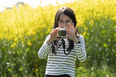 Uzun saçlı, gözlüklü ve çizgili tişörtlü güzel bir kız baharda sarı kolza tohumu tarlasının önünde fotoğraf çekiyor. Çocukların yaratıcılığı ve rahatlığı kavramı