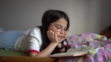 Beyaz tişörtlü ve gözlüklü güzel bir genç kızın çocuk odasındaki yatakta uzanmış kitap okuduğu bir video. Eğitim kavramı