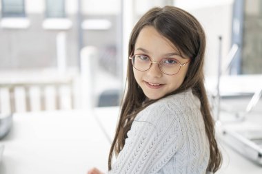 Gözlüklü ve svetşörtlü sevimli bir kız. Pencere arkasından kameraya dönmüş gibi görünüyor. Güzel bir oyunculuk çocuğu kavramı