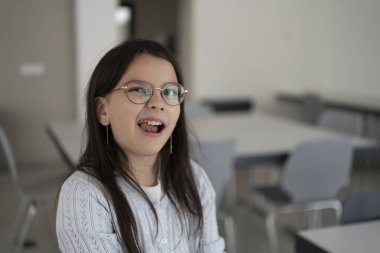 Gözlüklü ve kazaklı hoş bir kız okul kafeteryasındaki bulanık arka planda dururken titrek dişlere dille dokunuyor. Diş ve öğretici çocukların konsepti