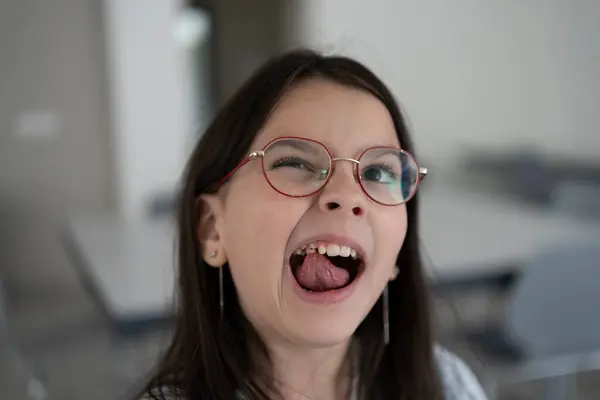 Gözlüklü ve kazaklı hoş bir kız okul kafeteryasındaki bulanık arka planda dururken titrek dişlere dille dokunuyor. Diş ve öğretici çocukların konsepti