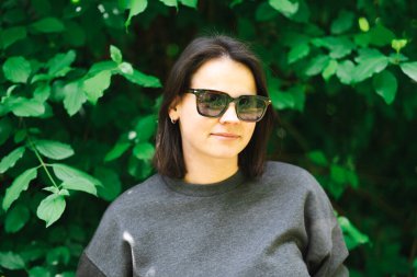 Yeşil yapraklara karşı güneş gözlüklü ve kazaklı gülümseyen genç kadın. Kavram: Doğal güzellik, kaygısız neşe, açık hava yaşam tarzı, yeni bir başlangıç, gündelik moda.