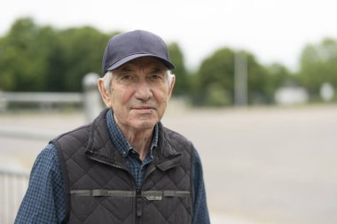 Baharda açık havada duran beyzbol şapkalı ve yelekli yaşlı bir adam. Kavram: aktif yaşlanma, mevsimsel tazelik, açık hava neşesi, canlılık.