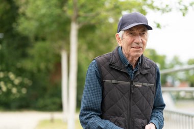 Baharda açık havada duran beyzbol şapkalı ve yelekli yaşlı bir adam. Kavram: aktif yaşlanma, mevsimsel tazelik, açık hava neşesi, canlılık.