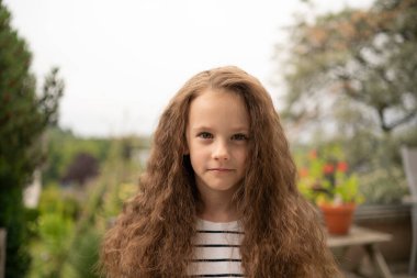 Çiçeklerin arasında çizgili tişörtlü, kıvırcık saçlı üzgün kızın yakın çekimi. Kavram: çocukluk duyguları, sessiz keder, doğa ve duygu, içsel an.