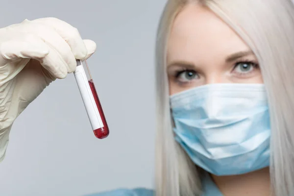 stock image young woman wearing a medical face mask and medical gloves is handling a blood probe in front of a grey background