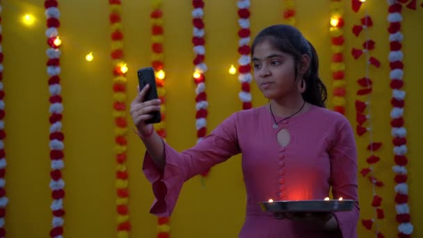 美しいと若い女の子は妖精の光の背景で彼女の手にDiwali油Diyaを保持し スマートフォンから自分自身を取ります ディワリ光の祭りは 女の子を楽しんで — ストック動画