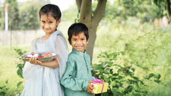 Hindu Brother and sister in ethnic wear holding Indian sweets and gift box on the occasion of Raksha Bandhan festival