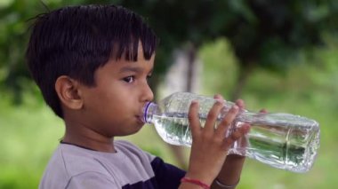Bahçede sıcak bir günde plastik şişe tutan ve su içen küçük Hintli çocuk konsepti..