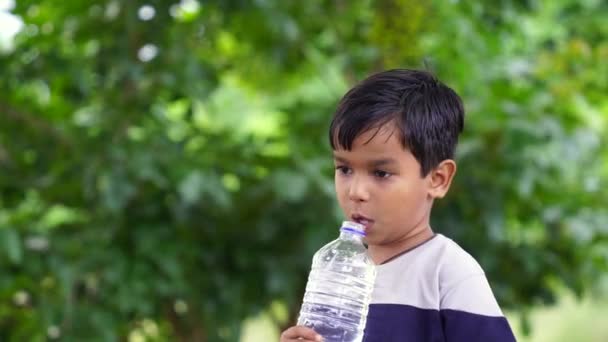 公園で黒髪の飲料水を飲んだハッピーかわいいインディアン少年 プラスチックボトルを持って親指を現す アウトドアショット — ストック動画