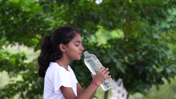 公園で黒髪の飲料水を飲んだハッピーかわいいインディアンガール プラスチックボトルを持って親指を現す アウトドアショット — ストック動画